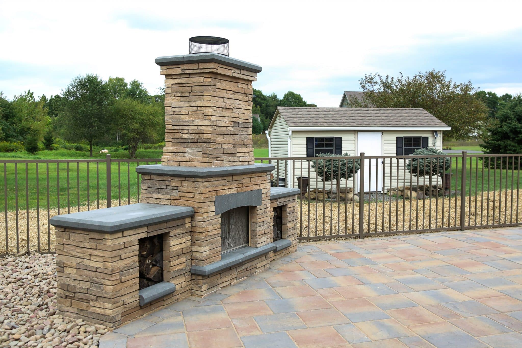 A Belgard Bordeaux series outdoor free standing fireplace with side wood boxes on a Belgard patio with pool fencing.