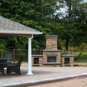 A free-standing precast concrete Belgard fireplace with side woodboxes on a pool patio in front of a fence next to a cabana.