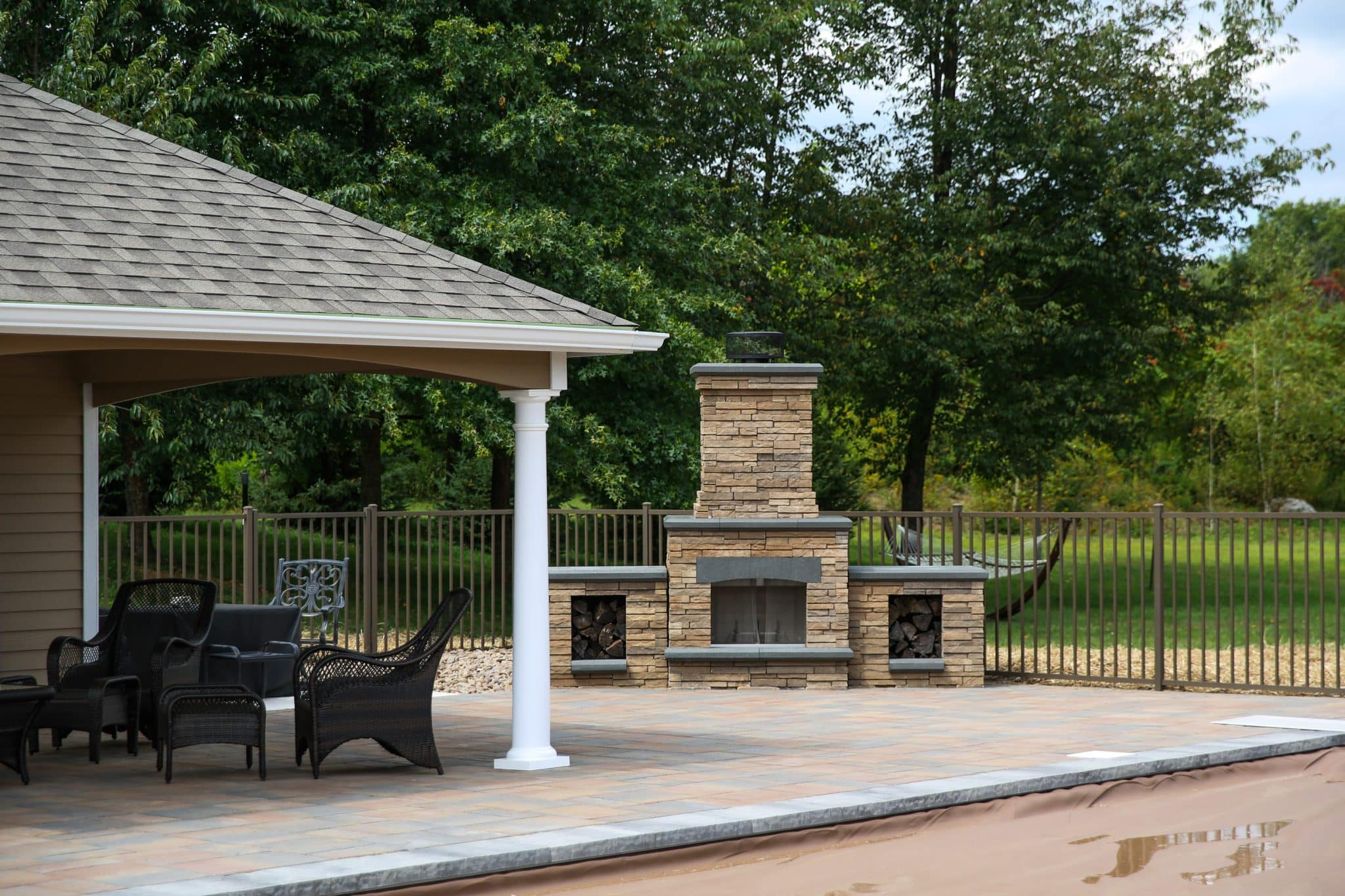 A free-standing precast concrete Belgard fireplace with side woodboxes on a pool patio in front of a fence next to a cabana.
