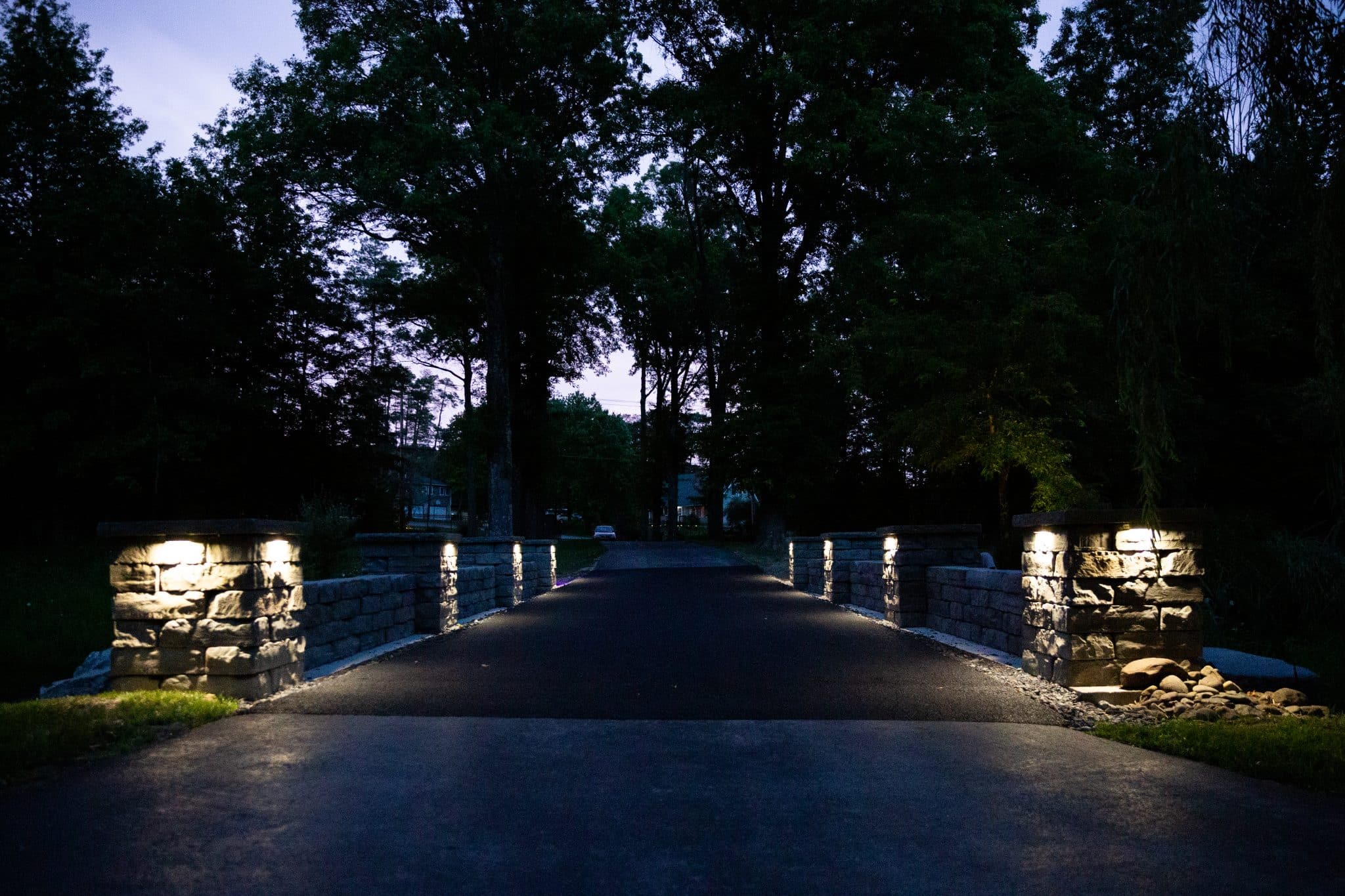 A head-on view of a driveway at night with two precast concrete stone texture retaining walls with pillars on either side. The scene is lit by outdoor lighting installed on each pillar. Designed and installed by Masseo Landscape, Inc, New Paltz Landscapers.