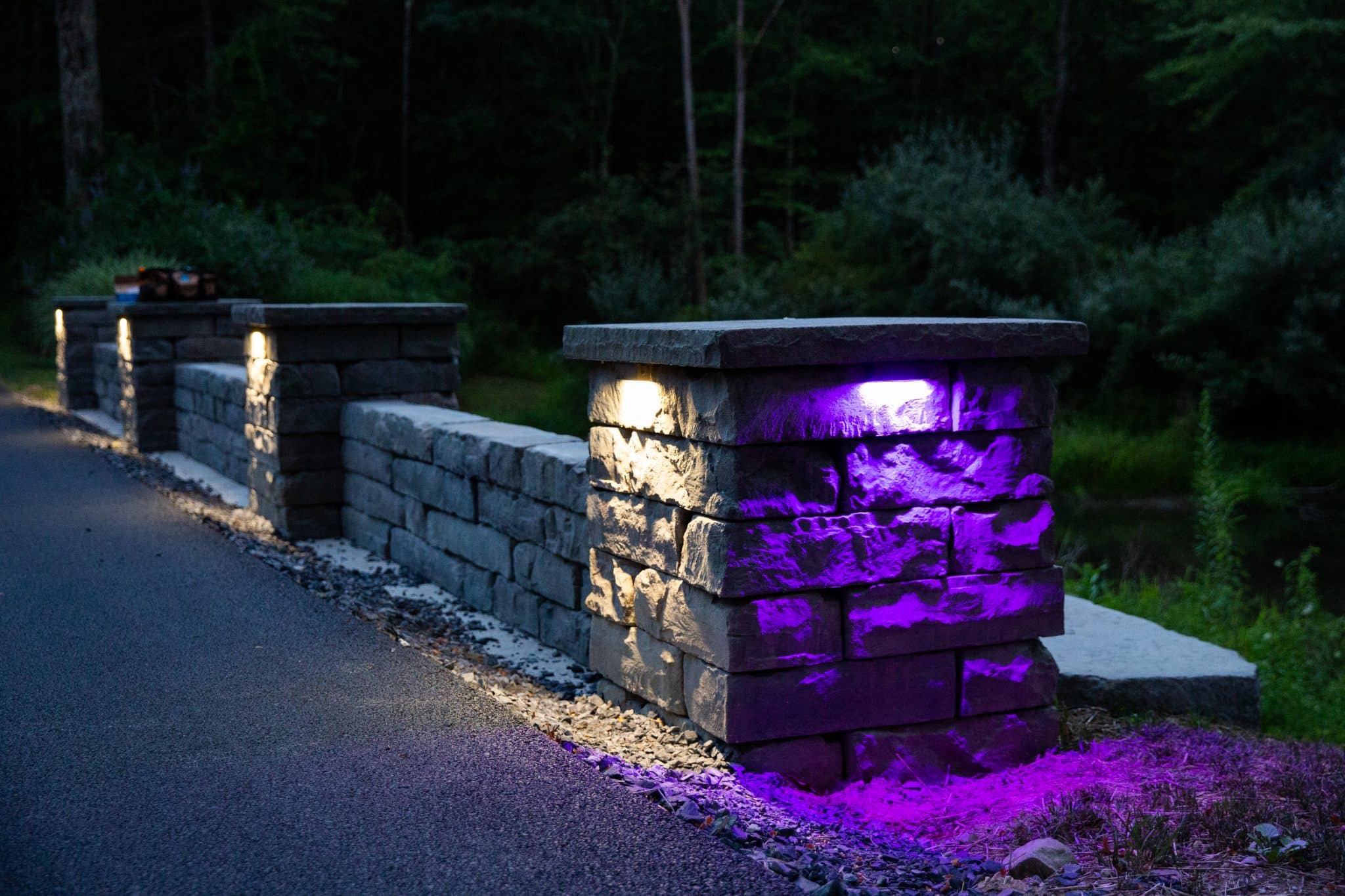 A purple light illuminates a stone-texture precast concrete pillar set into a free standing wall along a driveway. Alliance color-changing LED outdoor lighting and Rosetta Kodah wall installed by Masseo Landscape, Inc.