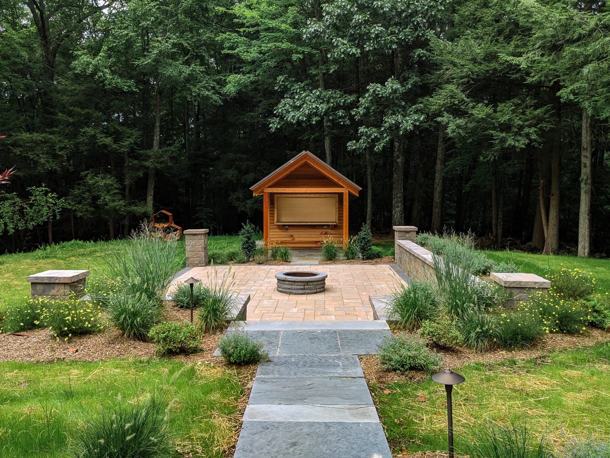 A wooden bar shed with an overhang behind a tan precast concrete Belgard paver patio with a round fire pit. A bluestone walkway, native plantings, and outdoor lighting fixtures are in the foreground. Designed and installed by Ulster County Landscapers, Masseo Landscape, Inc.