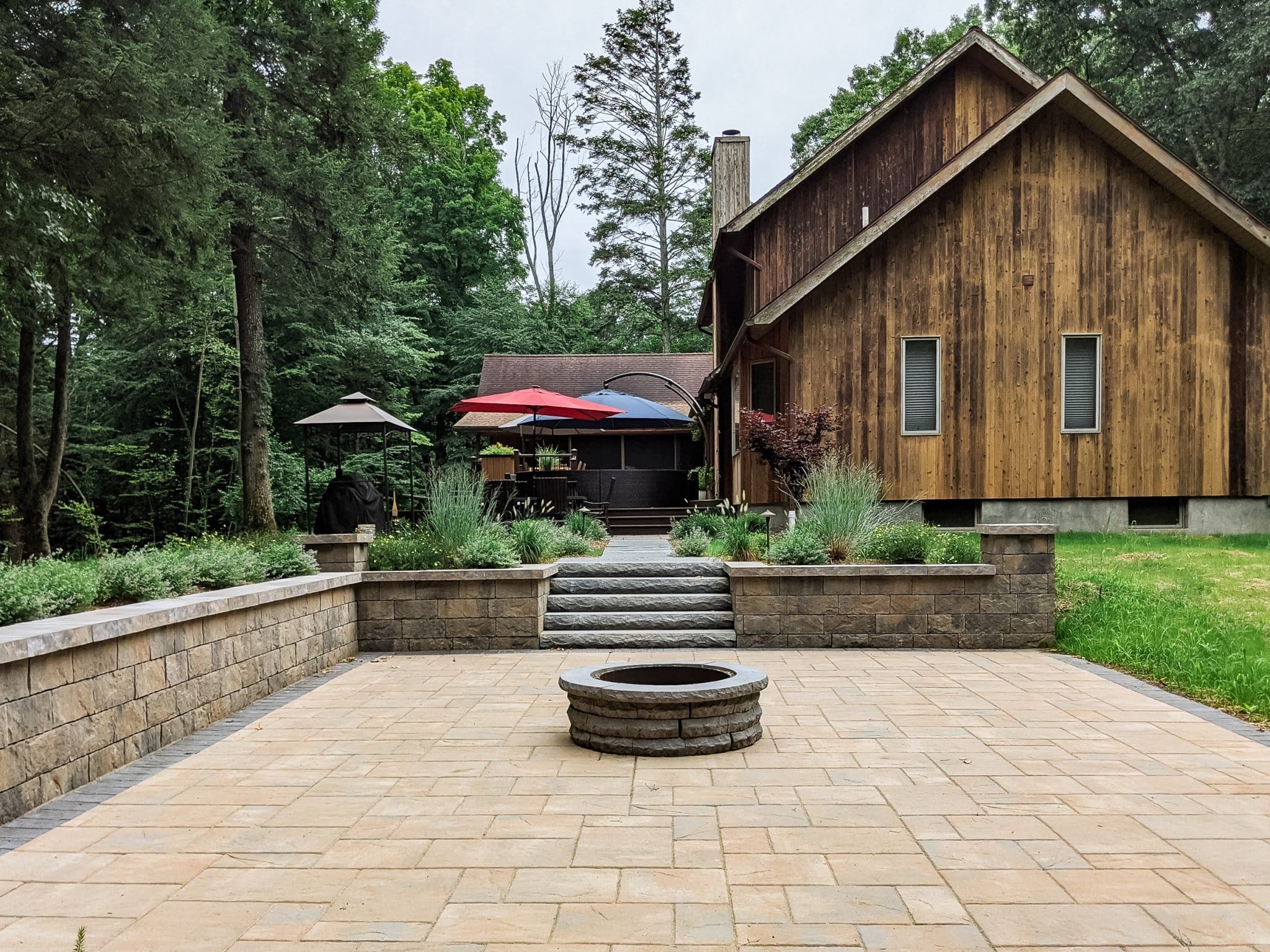 A tan precast concrete paver patio, retaining walls, and steps with a circular fire pit and contrasting paver border next to a wooden house in Stone Ridge, NY by Masseo Landscape, Inc.