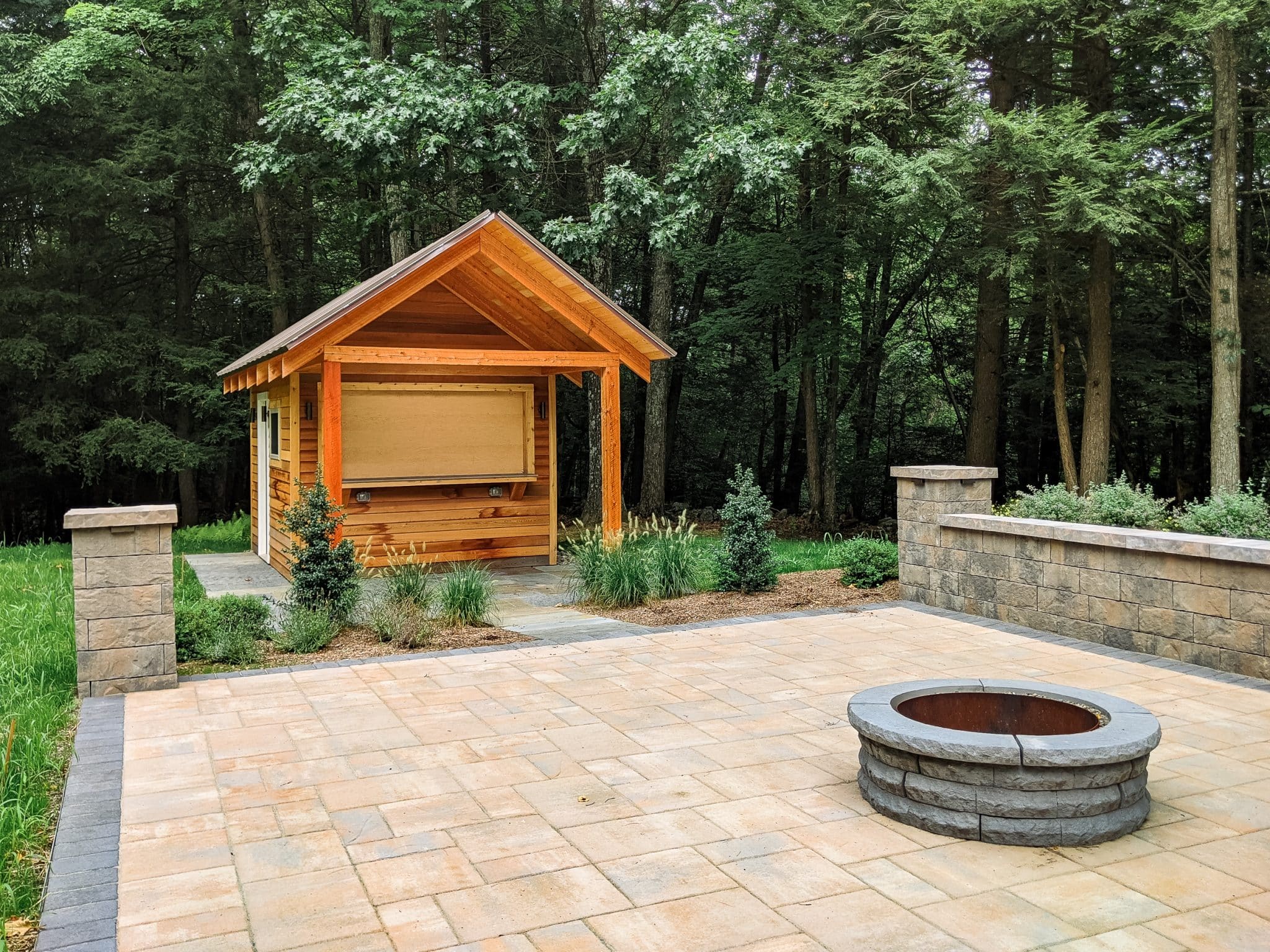 A wooden bar shed next to a precast concrete patio with pillars and a retaining wall and a circular firepit, installed by Masseo Landscape, in Stone Ridge, NY