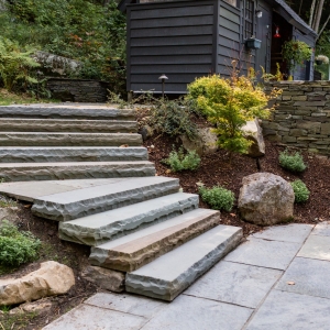 Stone face bluestone staircase set into a hillside with catmint and Japanese maple plantings set with natural boulders and landscape lighting next to a bluestone pool patio in Woodstock, NY.