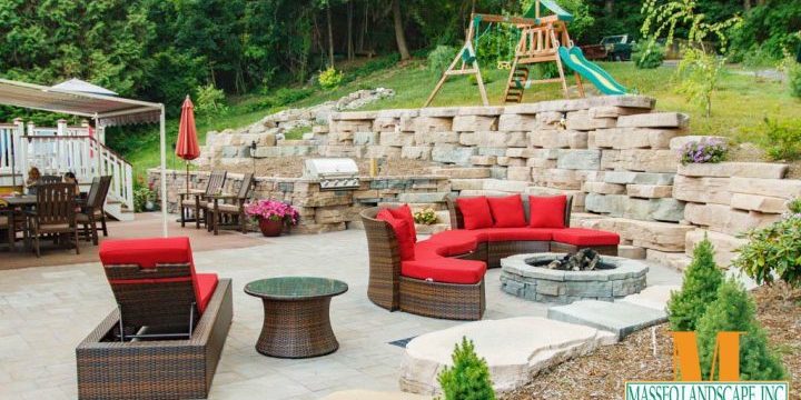 An outdoor living area with a Cambridge Pavers patio, retaining walls built from Rosetta Hardscapes wall stone, a circular fire pit, grill station, and built in seating.
