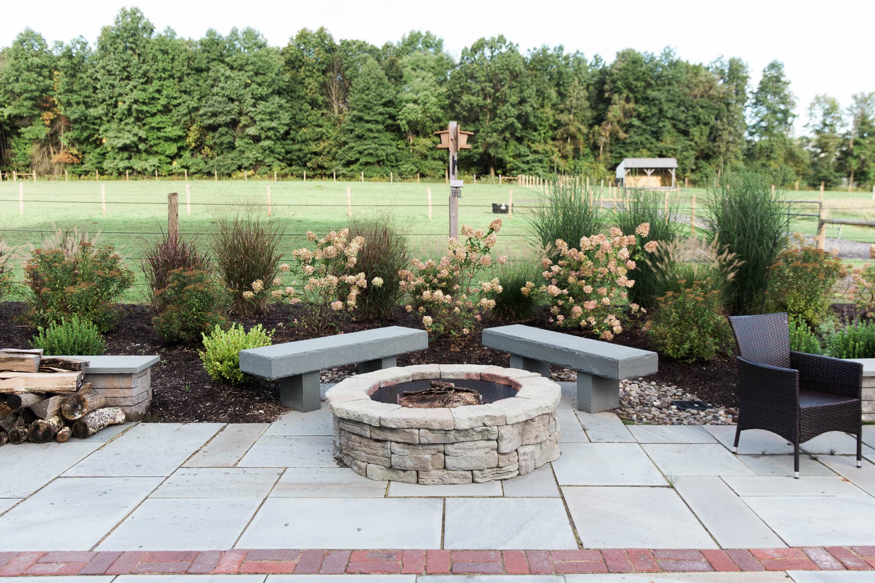 A circular precast concrete fire pit and bluestone slab benches on a rectilinear bluestone patio with native plants like hydrangea and spirea behind, installed by Masseo Landscape, Inc. Hudson Valley Landscaper