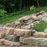 A custom water feature built with a blend of precast concrete and natural stone, with ambient outdoor lighting, installed and designed by Masseo Landscape, Inc.`